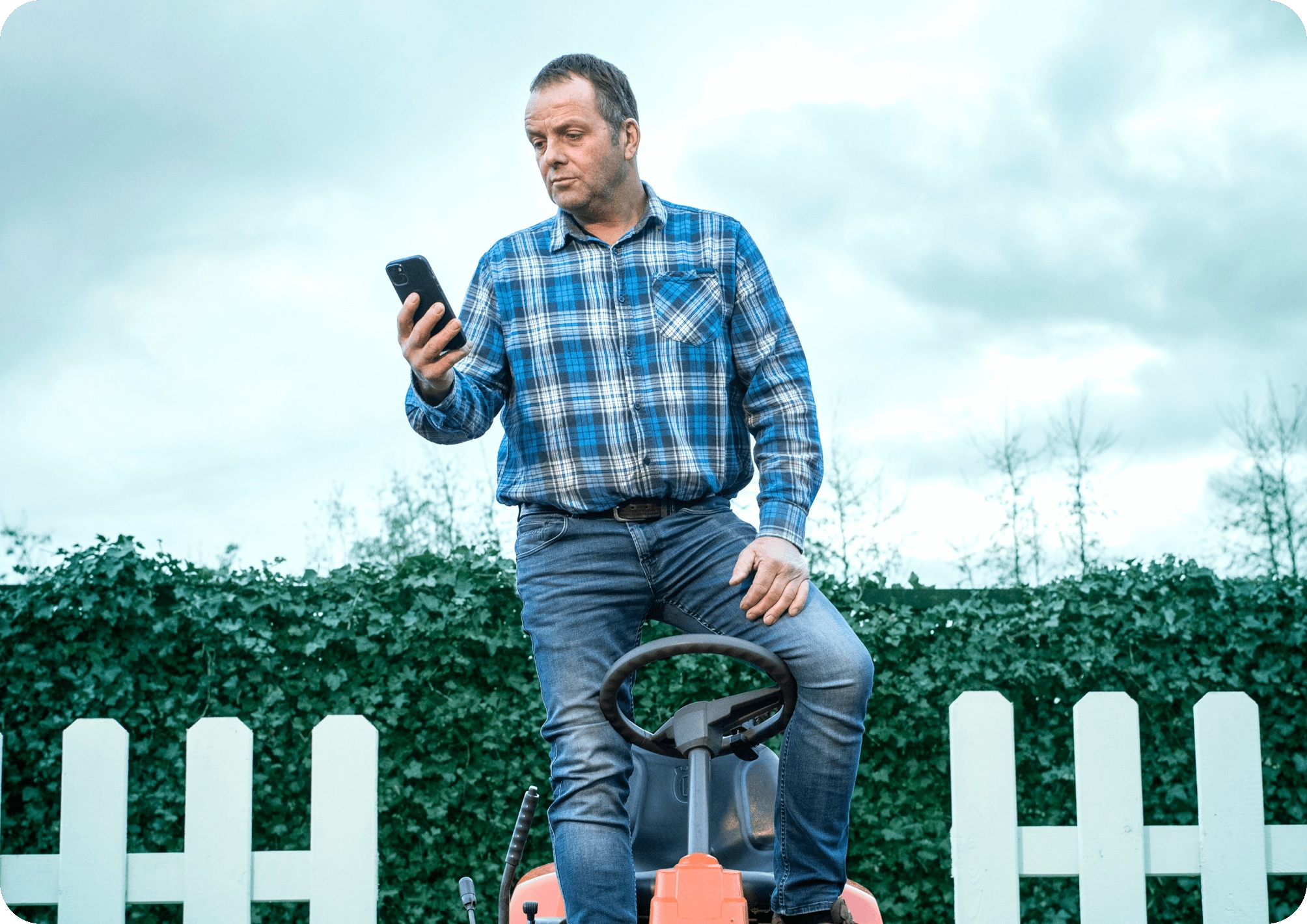 Hombre de pie en tractor cortacésped al aire libre, mirando smartphone bajo cielo nublado.